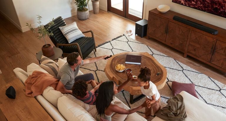 A family sitting in a room. A man controls a sound bar with a remote.